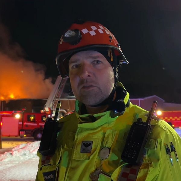 En brandman i röd hjälm och gul uniform står framför en brinnande byggnad och flera räddningsfordon.