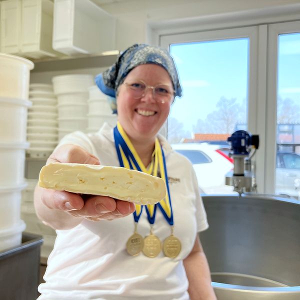 Person håller ostbit i handen, bär medaljer. Osttillverkning pågår i bakgrunden.