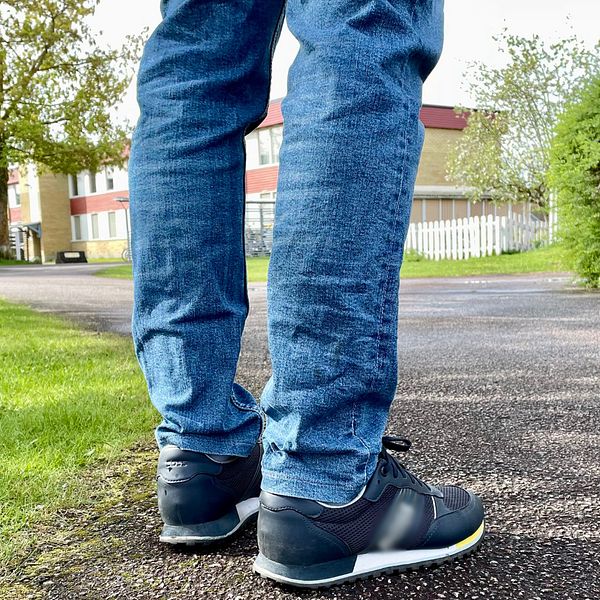 Person i jeans och sneakers står på en asfalterad gångväg i grönskande parkmiljö.