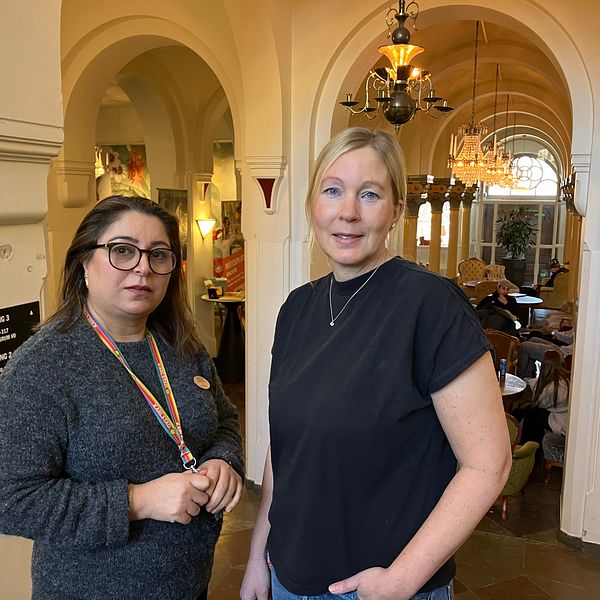 Två personer står i en elegant hall med valv och hängande lampor i bakgrunden.