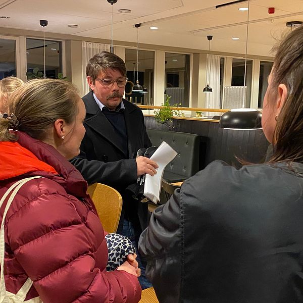 Tre personer samtalar i en inbjudande restaurangmiljö. En kvinna i en röd jacka vänder sig mot en man med dokument i handen, medan en annan kvinna står i bakgrunden. Belysningen i rummet är mjuk och det finns växter på bordet som ger en hemtrevlig atmosfär.