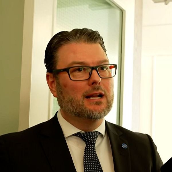 Edward Riedl (M) i kostym intervjuas av flera journalister som håller fram mikrofoner.