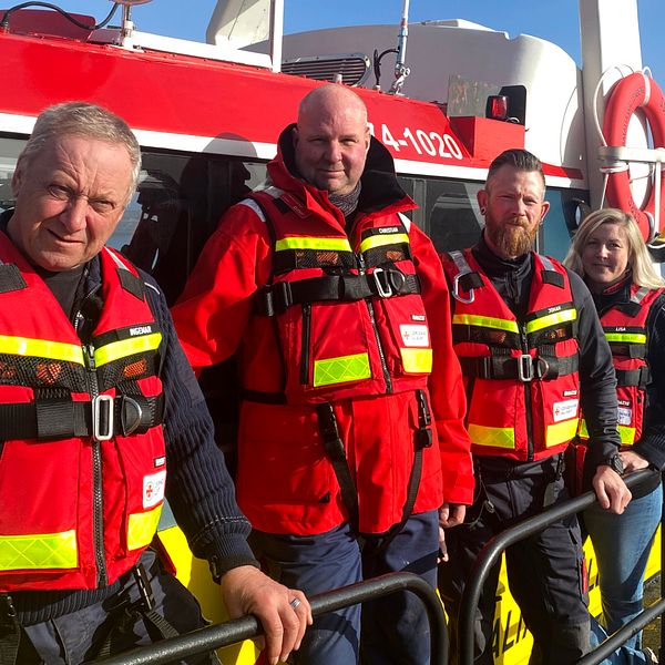 Fyra sjöräddare i räddningsdräkter står på däck vid en röd räddningsbåt.