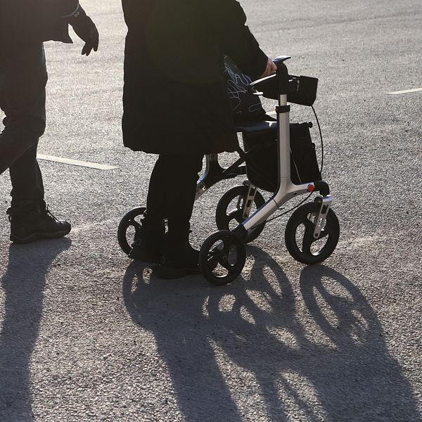 Halvbild på två äldre personer på en parkering. Mannen går själv och kvinnan har rullator.