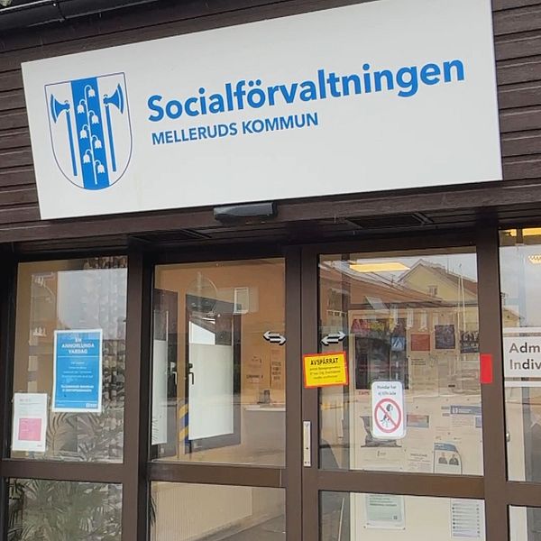 Entrén till Socialförvaltningen i Melleruds kommun, med en skylt som tydligt visar namnet. Flera informationsskyltar är synliga vid dörren, och en polisbil finns parkerad i bakgrunden.