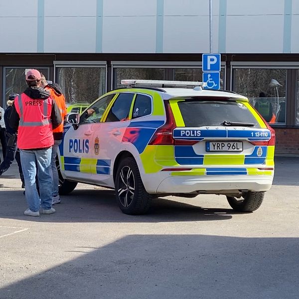 Polisbil parkerad utanför en byggnad. Person i röd väst pratar med flera människor.