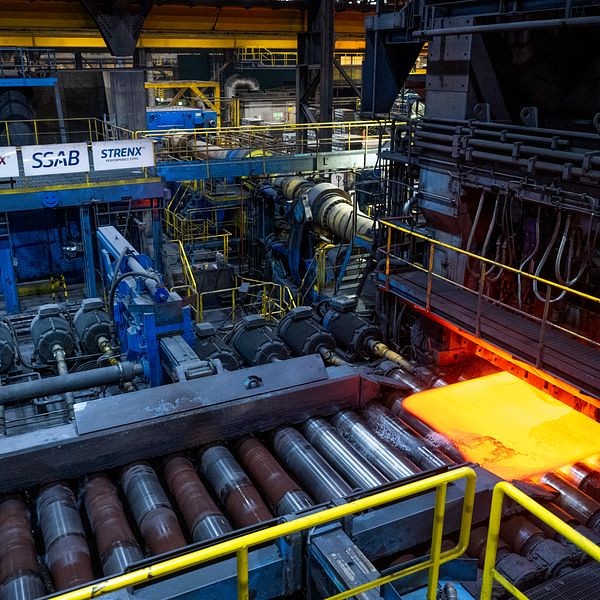 Rullande stålplatta i en industriell produktionslinje, omgiven av maskiner och utrustning. Plattan är lysande orange, vilket indikerar att den är uppvärmd. Skyltar med varumärken syns i bakgrunden tillsammans med en varningsskylt längst ner.