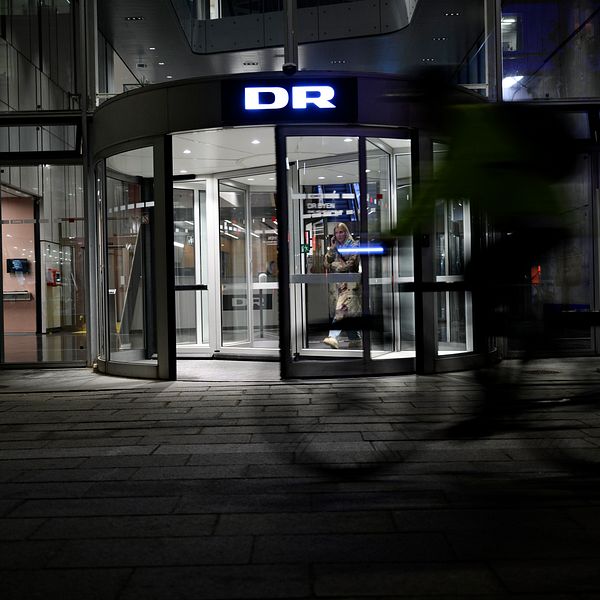 Entrén till DR:s byggnad i skymningen, oskarp cyklist i förgrunden.