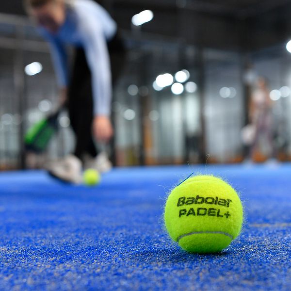 En padelboll ligger på ett padelbanans blå matta, suddig spelare i bakgrunden.