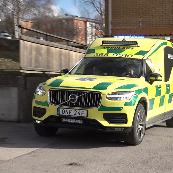 En ambulans i gul och grön färgsättning kör ut från en garageport i ett urbant område, med träddelar och byggnader i bakgrunden.