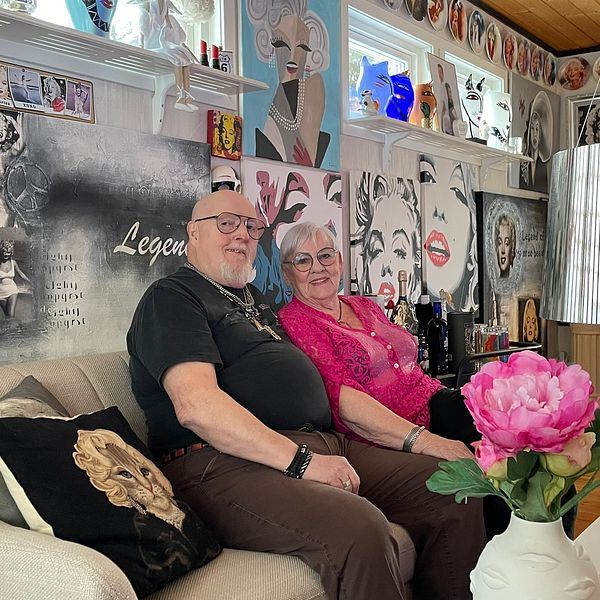 Ett par sitter på en beige soffa i ett inrett rum med en konstnärlig vägg prydd med bilder och citat. Alla bilder föreställer filmstjärnan Marilyn Monroe. Rummet har en modern taklampa och en blomma i en vas på bordet.