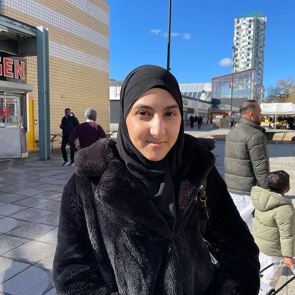 En ung kvinna står på Tensta torg i svart jacka och ler. Det går folk i bakgrunden.