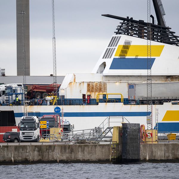 Färja vid kajen med vit och blå-gul randig överdel på ett varv, omgiven av industriell utrustning och lastbilar i en hamnmiljö.