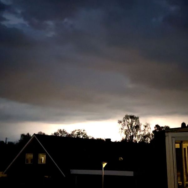 Mörka regnmoln med kraftiga blixtar över villaområde i skymning med siluetter av träd och hus mot horisonten.