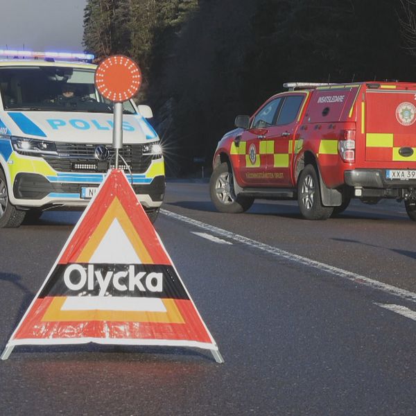 En polisbil och en räddningstjänstbil står på en väg som är avstängd med varningsskyltar som visar texten ”Olycka”. Varningsskyltarna är placerade på körbanan för att informera trafikanter om en incident. I bakgrunden syns träd och berg.