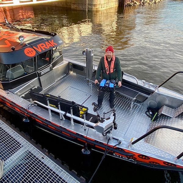 En ca 10 meter lång plåtbåt med styrhytt och stora öppna däck. Båten är uppmärkt med stora orange bokstäver ”VRAK”  – museet. PÅ däcket står en man med en undervattensdrönare i händerna. det ser ut som en liten u-båt.