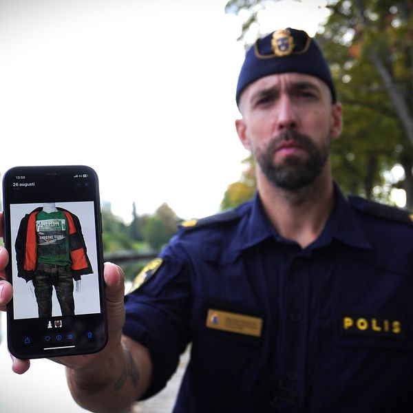 En polis visar en bild av en jacka på sin mobiltelefon. Bakgrunden inkluderar en vattenspegel och träd. Jackan är i färgerna röd och svart och bärs av en person på bilden.