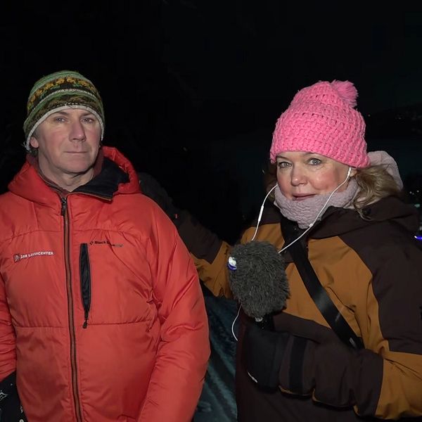 En person i röd jacka står bredvid en person i mörkare kläder och en rosa mössa, medan de pratar i ett snölandskap under natten. Staden syns svagt i bakgrunden, belyst av ljus.