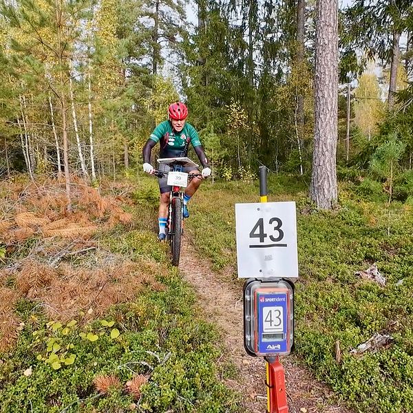 En cyklist på en stig i en skog passerar en skylt med nummer 43. Omgivningen består av grönskande buskar och träd, med höstliknande färger i markvegetationen.