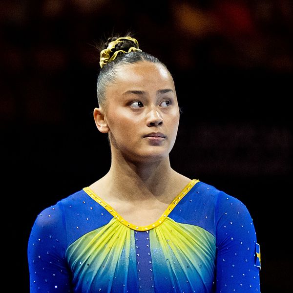 En gymnast i en färgglad, glittrande dräkt med blå och gula detaljer står på scenen under en tävling. Bakgrunden visar en oskarp åskådararena, vilket ger en känsla av en spännande sporthändelse.