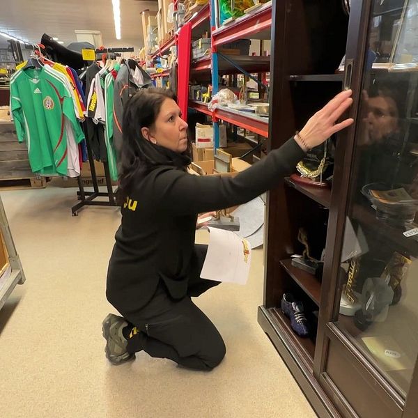 Kvinna i lokal organiserar hyllor med glasdörrar, omgiven av kläder och varor från auktionen av Svennis kvarlåtenskap.