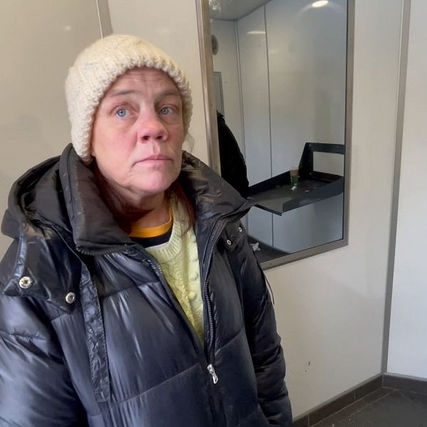 Petra Källberg med en mörkgrå täckjacka och vit stickad mössa är hemlös. Hon visar upp en offentlig toalett där hon brukar sova, väggarna är vita och det är en spegel i bakgrunden.