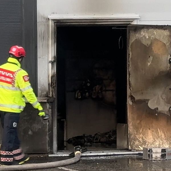 Två brandmän i gul och svart skyddsutrustning står vid en brinnande dörröppning till en byggnad. Dörren är delvis öppen och visar tecken på brandskador, med svart sot på väggarna inuti. En brandslang ligger på marken framför dem, vilket indikerar pågående släckningsarbete.