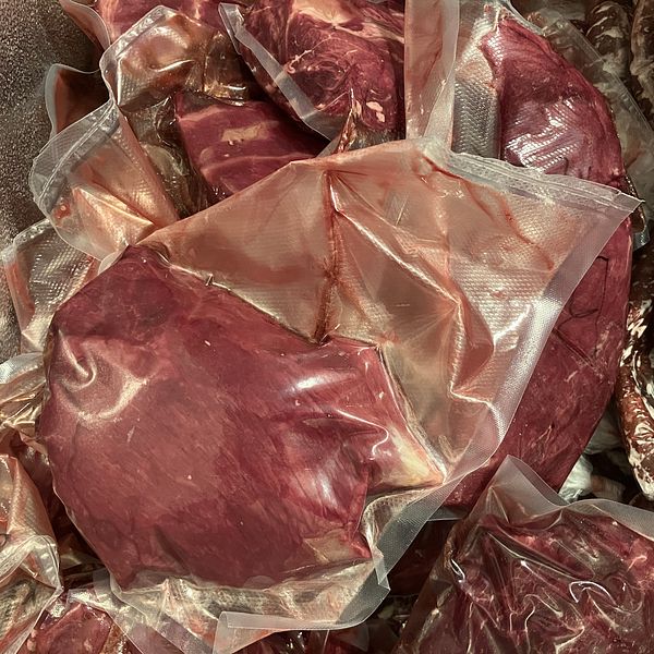 Förpackat kött i vakuumförpackningar, staplat i en frys eller kyl. Köttet har en mörkröd färg och är synligt i flera olika förpackningar.