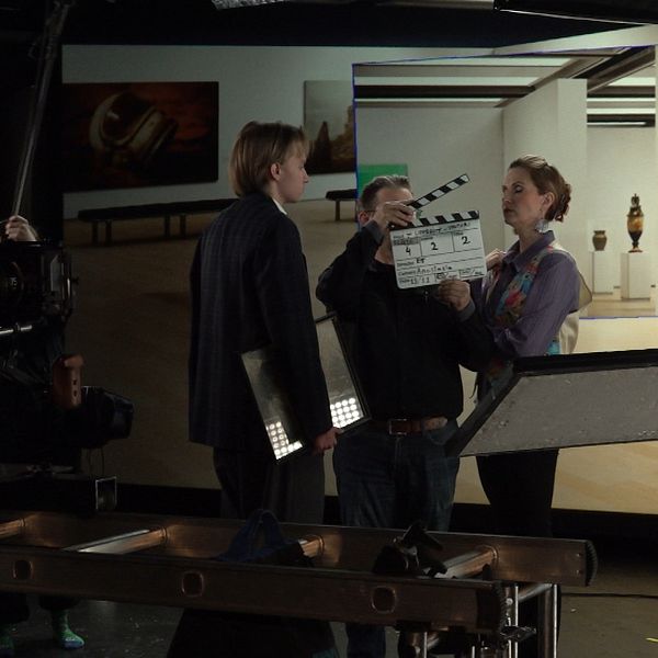 En filmproduktion pågår inomhus, där en person står framför kameran och förbereder sig för en scen. En annan person håller en klappa (slate) medan två personer diskuterar i bakgrunden. I rummet syns en konstutställning med olika verk på väggarna, vilket ger en avslappnad och kreativ atmosfär. Kamerautrustning syns vid sidan och belysning används för att belysa scenen.