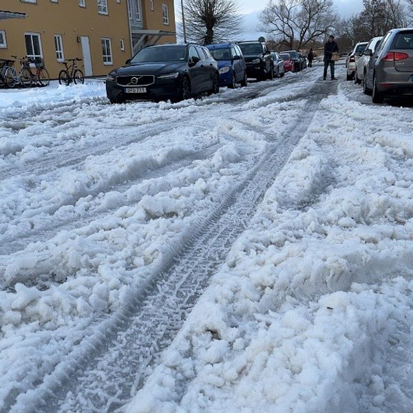 En snötäckt gata med djupt snö spår efter bilar. Parkerade bilar syns på båda sidor om vägen, och en person går på den snöiga trottoaren.