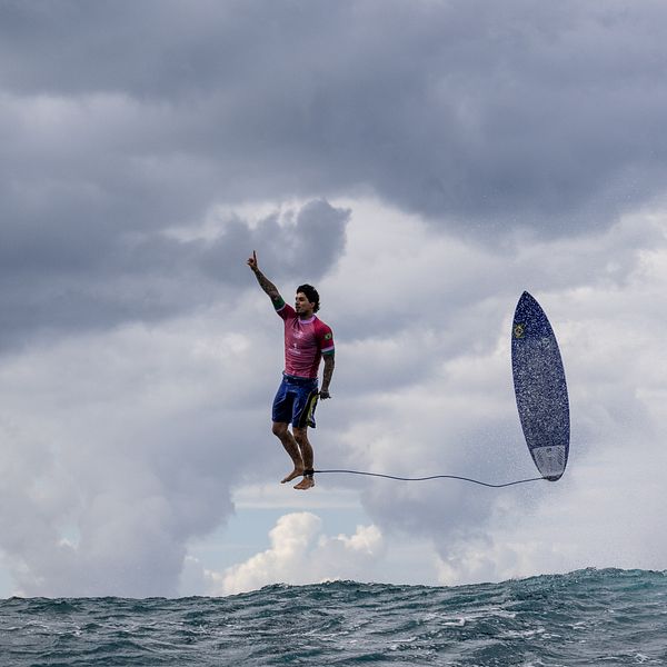 Surfare i rosa tröja hoppar över vattnet med en surfbräda i handen, omgiven av mörka moln och en stormig himmel.