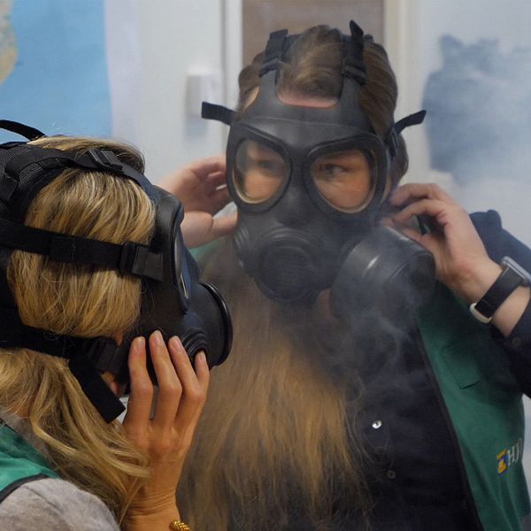 Två personer står i ett rum och justerar sina gasmasker medan röken sprider sig kring dem. Båda bär gröna västar och syns i en utbildnings- eller träningstillställning som handlar om säkerhet eller krisberedskap. En karta kan ses i bakgrunden, vilket tyder på att platsen är inomhus.