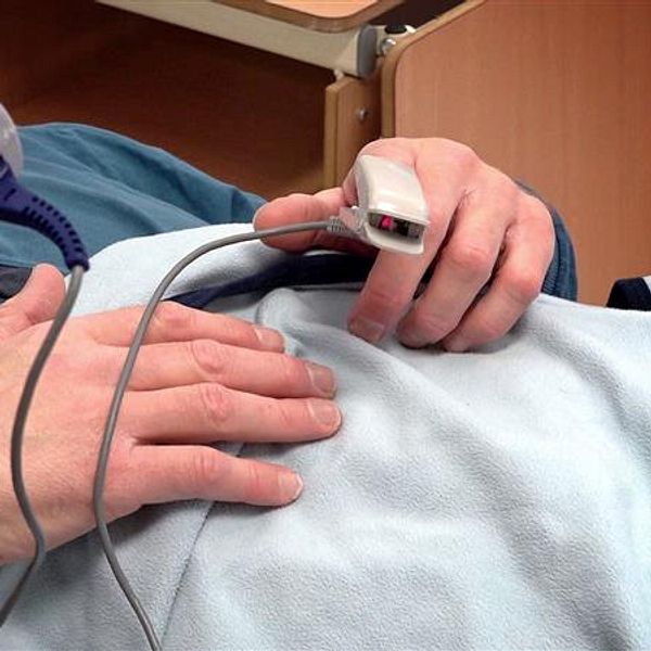En person i sjukhussäng mäter syremättnad med pulsoximeter på fingret.