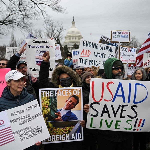 En grupp människor står samlade framför Capitolium i Washington D.C. och håller upp skyltar som stöder USAID, där budskap som ”USAID SAVES LIVES” och ”THE FACES OF FOREIGN AID” är framträdande. Många deltagare håller också amerikanska flaggor, och stämningen i bilden tyder på en aktiv demonstration för biståndsmyndigheten.