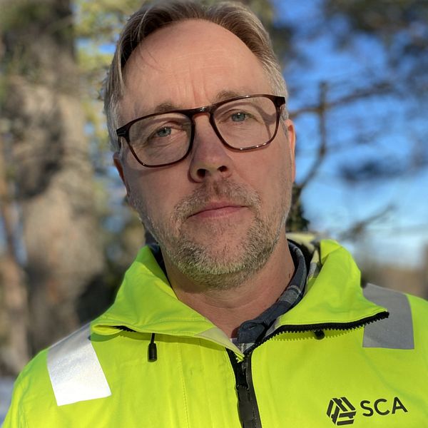 Person i gul SCA-jacka framför skogsmiljö under en solig dag.
