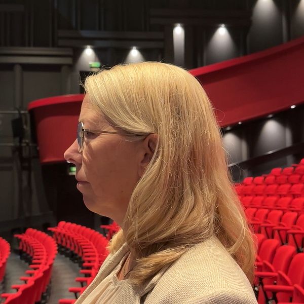 En person med långt blont hår står i profil i en teater. I bakgrunden syns ett stort antal röda stolar i publikområdet, med en mörk scenyta till vänster.
