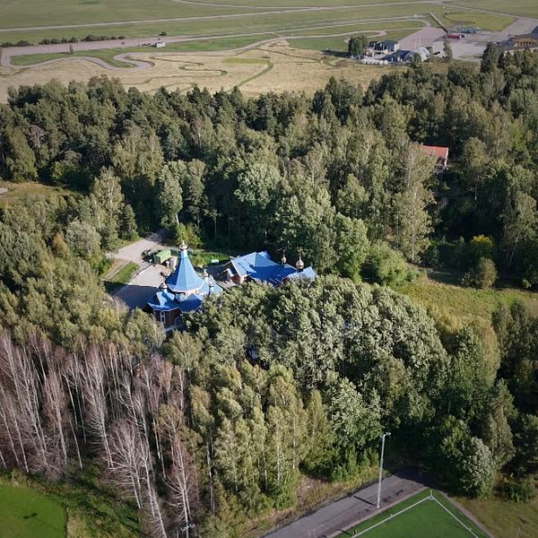 Flygfoto över ett hus med blått tak omgivet av tät skog och öppna fält.