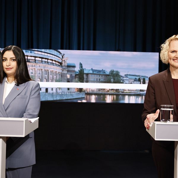 Två politiker står vid talarpodium under en debatt med riksdagsbakgrund på skärmen.