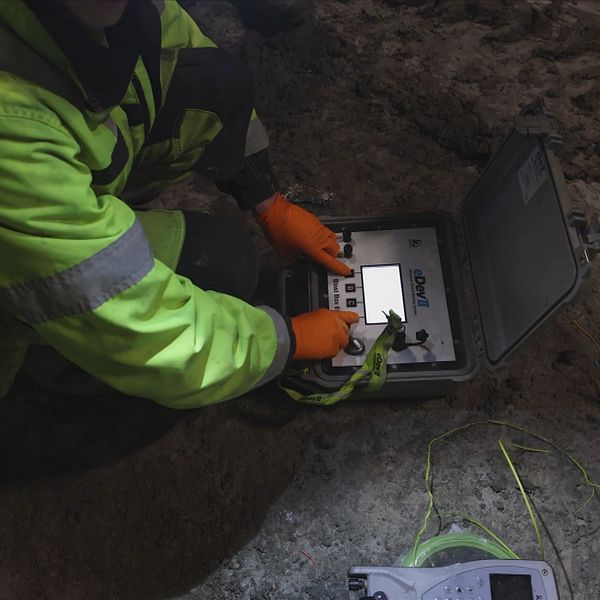 En person i skyddskläder med lysande gul jacka sitter på huk på en jordig mark och hanterar en teknisk enhet i en portabel låda, omgiven av sladdar och en elektronisk mätinstrument.