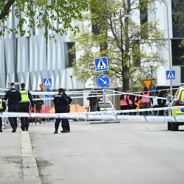 Poliser vid avspärrning och polisbil på gata utanför en modern byggnad.