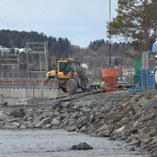 Grävmaskin arbetar vid vattnet nära en industriell anläggning och stenkantad strand.