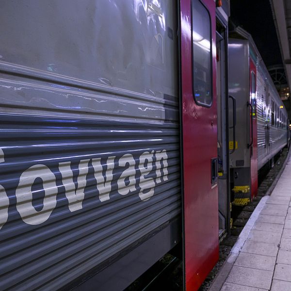 ALT-text: Tåg med texten ”Sovvagn” på sidan, stående vid en perrong i mörker, med plattformsbelysning och skymtor av människor som väntar.