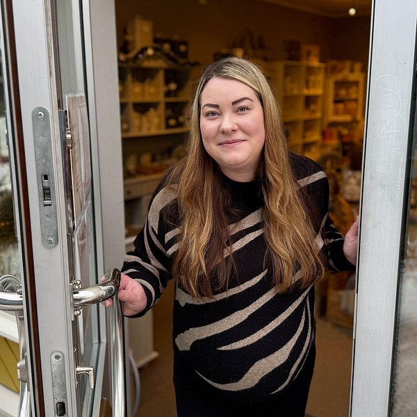 Jessika Skarin står vid ingången till sin butik i Orsa. Butiken är en presentbutik med inredning och presentartiklar. Det är vinter och snö syns på marken utanför.