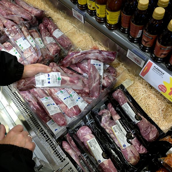 En person väljer bland olika sorters kött som ligger på ett kylskåp i en butik. I bakgrunden syns flaskor med sås och prisinformation för köttet.