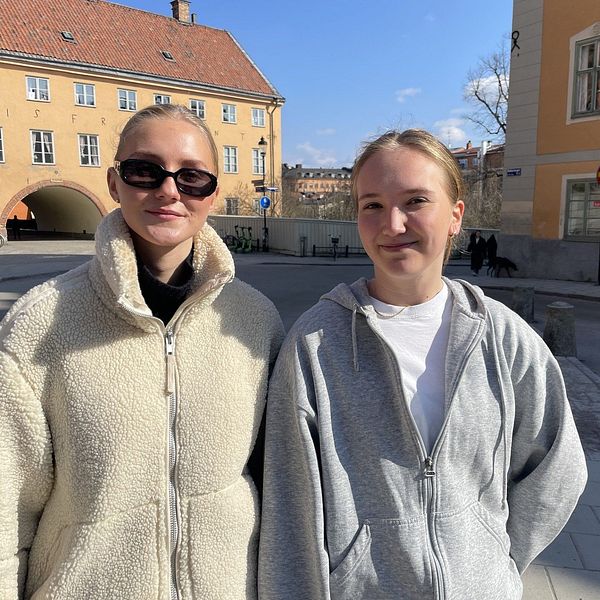 Två personer står utomhus i en solig stadsmiljö. Gamla byggnader i bakgrunden.