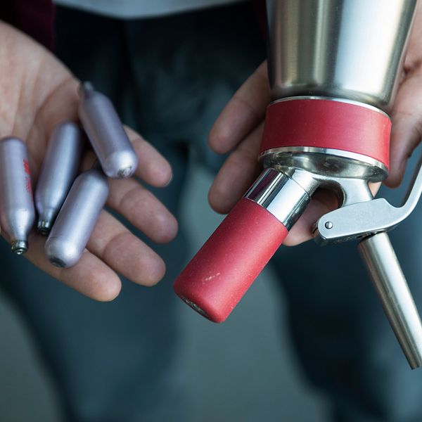 En person håller en metallpump med en röd spets i ena handen och flera silverfärgade gaspatroner i den andra handen.