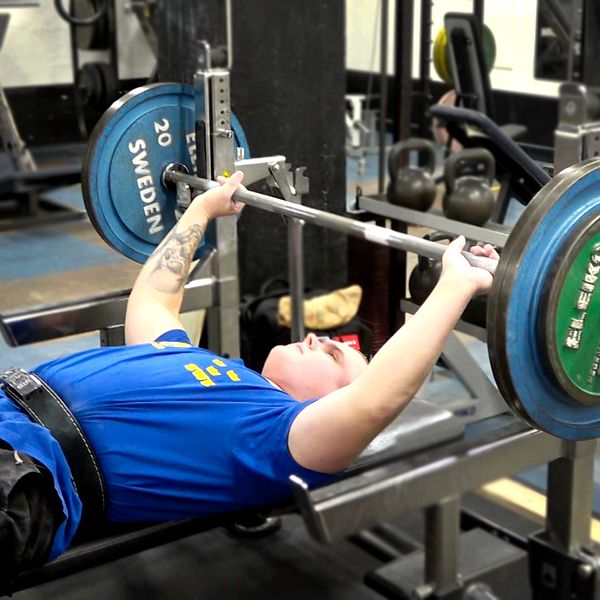 Simon Blom utför bänkpress i ett gym i Södertälje, liggande på en bänkbänk med en skivstång över bröstet. han bär en blå t-shirt med gul text och har ett bälte runt midjan för stöd. Vikterna på skivstången är markerade med färgade plattor. I bakgrunden syns andra träningsutrustningar och vikter.