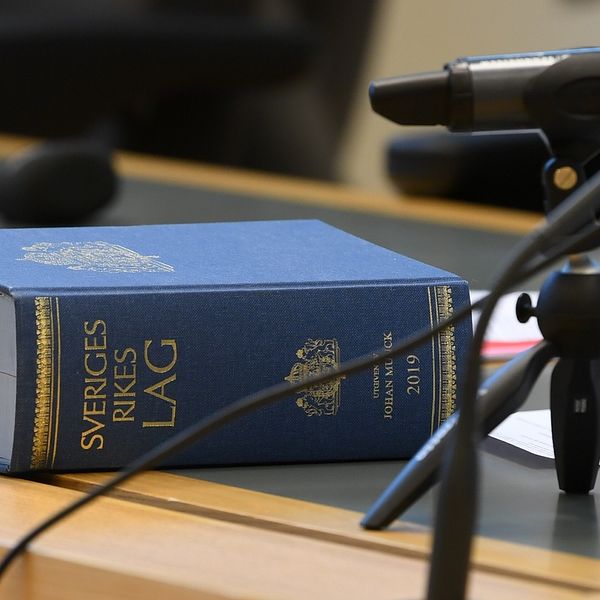 Sveriges Rikes Lag på ett bord i rättssal intill mikrofoner och anteckningar.