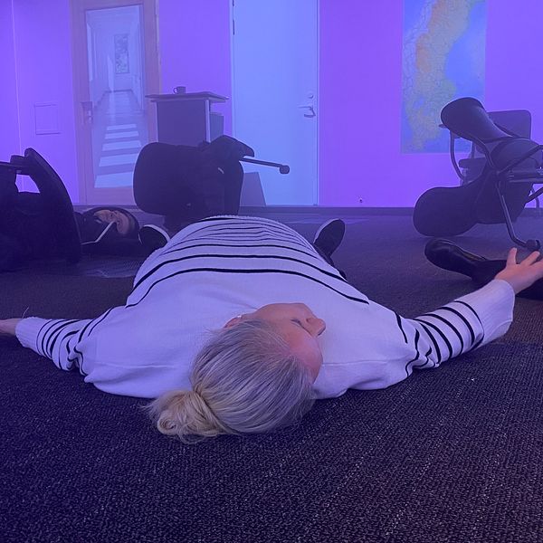 Kvinna liggande på golvet i ett kontorsmiljö med utspridda stolar och lila belysning, vilket kan indikera en simulering av en nödsituation eller träning i evakuering.