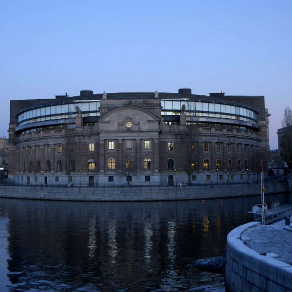 Svenkska riksdagshuset vid skymning, speglat i vattnet. Omgivningen visar en lugn stadsbild med belysning från byggnaderna i bakgrunden och en klar himmel.